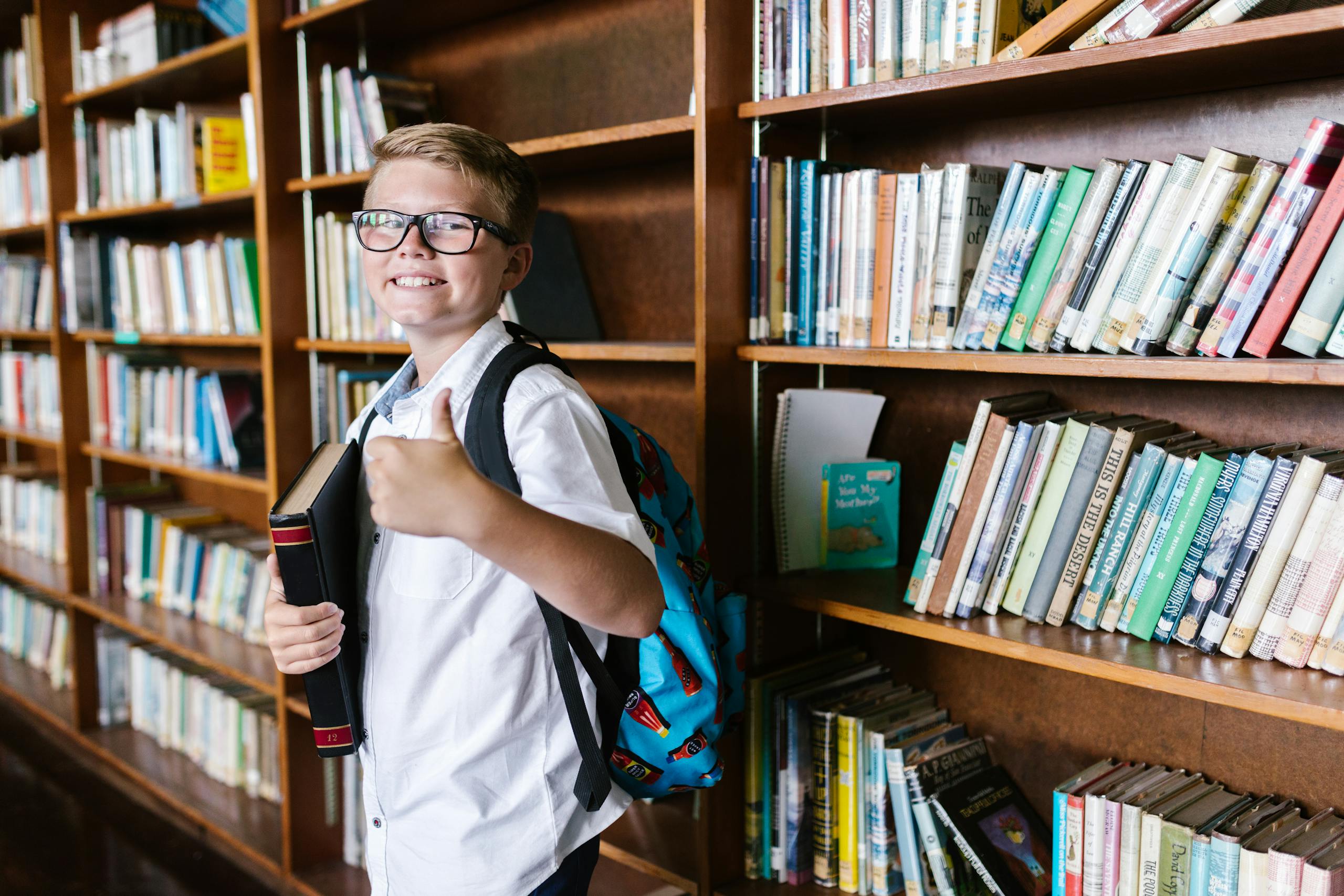 Young boy in a library gives a thumbs up while holding a book. Perfect for back-to-school themes.